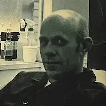 An old black-and-white photo of Jim Nicholson (Grandad), wearing a black button-down barber shirt with spray bottle and styling products arranged behind him.