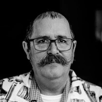 Black and white photo of Tim Collins, former owner of Tim's Barber Shop, wearing black rectangular glasses, a classic mustache with the ends curled up, a white barber shirt with vintage barber items illustrated, a checkered collared shirt underneath, and a white t-shirt beneath that